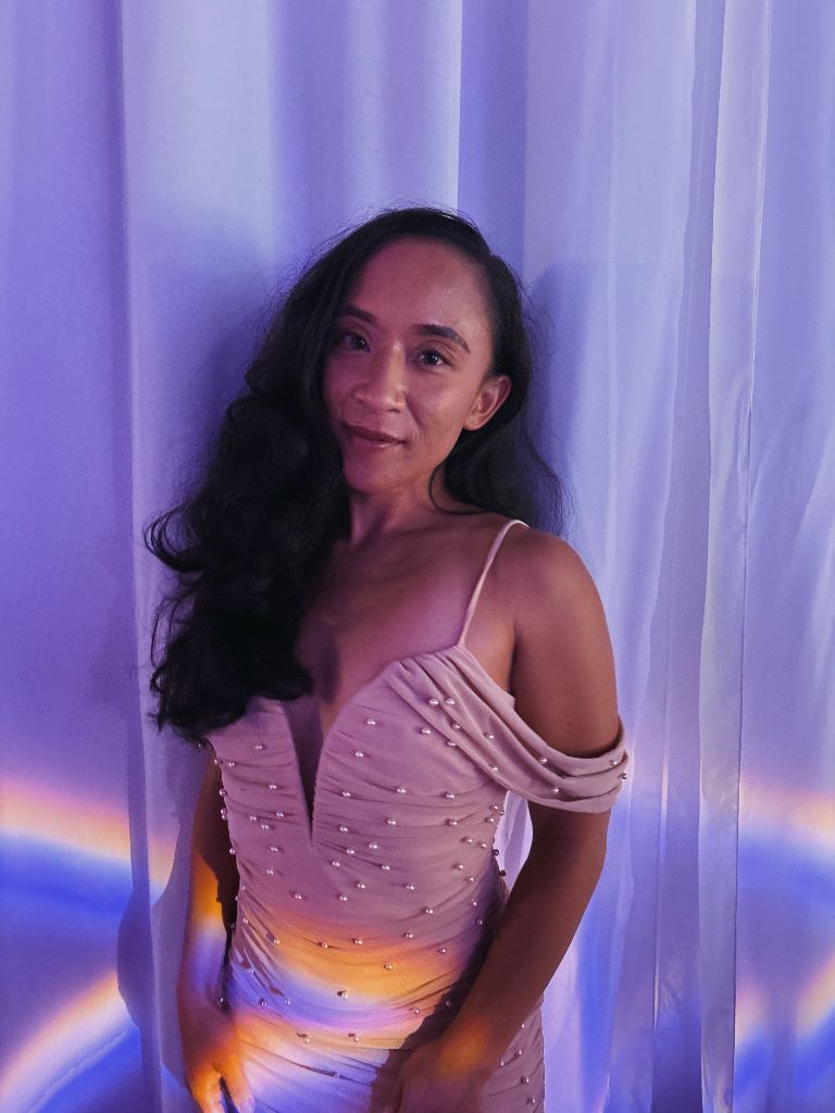 Professional headshot of Genel Ronquillo, Culture and Community Strategist for San Diego events business Sip & Social Club, posing in a pink pearl-embellished dress against a colorful curtain backdrop.