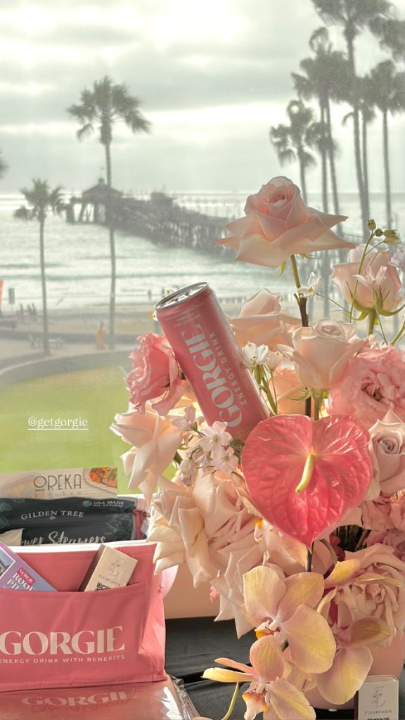 A pink Gorgie energy drink can nestled in light pink flowers on a rooftop with a pier and ocean view during a wellness event in San Diego. - Sip & Social Club San Diego event