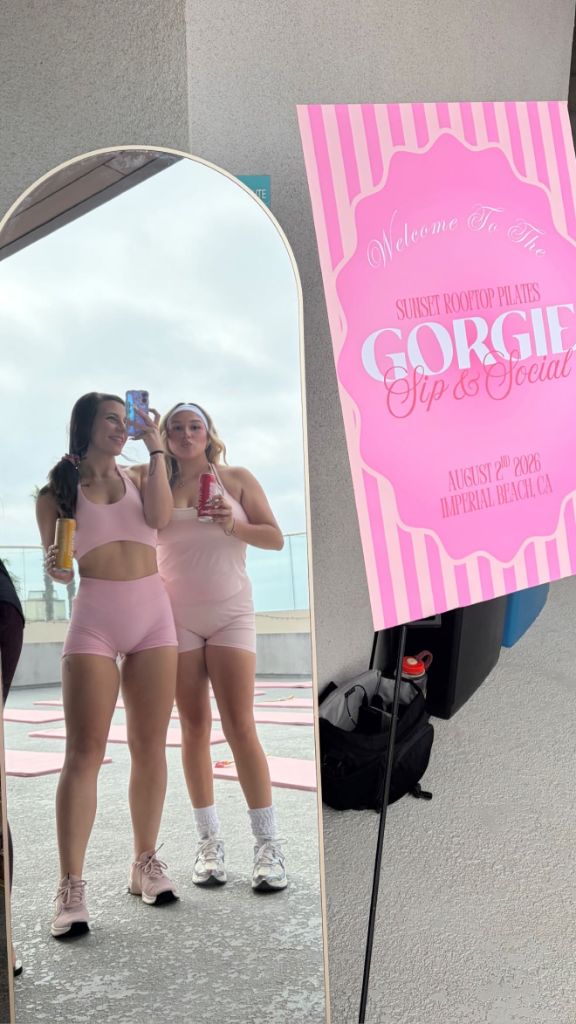 Two friends in pink activewear smiling for a mirror selfie next to a Gorgie Sip and Social event sign in Imperial Beach, California. - Sip & Social Club San Diego event