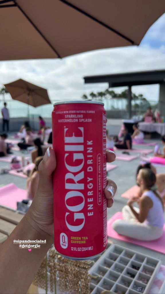 A close-up of a hand holding a Watermelon Splash Gorgie energy drink with a rooftop pilates class in the background in San Diego. - Sip & Social Club San Diego event