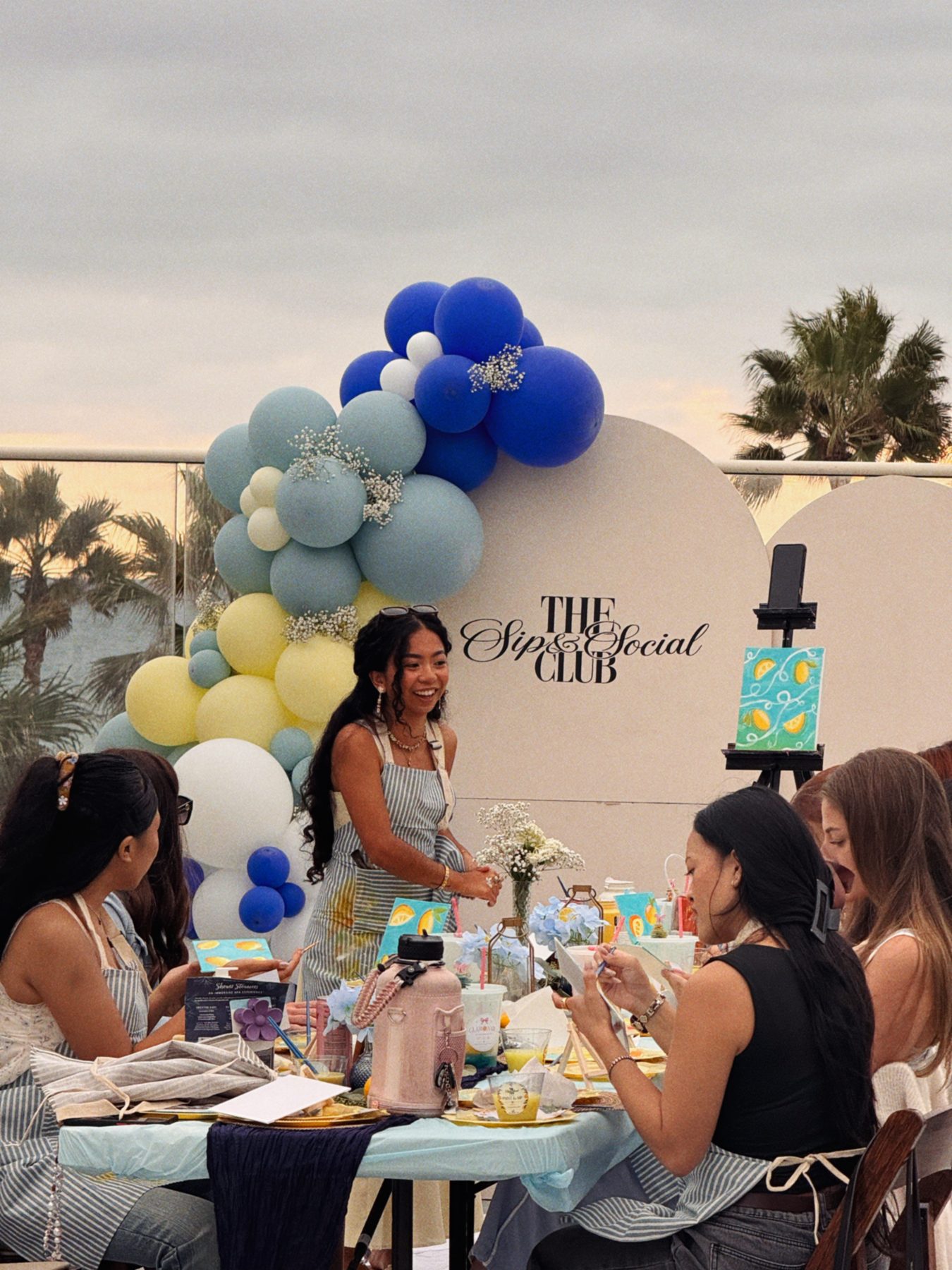 A smiling host leading a lemon-themed paint and sip at a long table in front of a balloon arch and The Sip and Social Club sign
