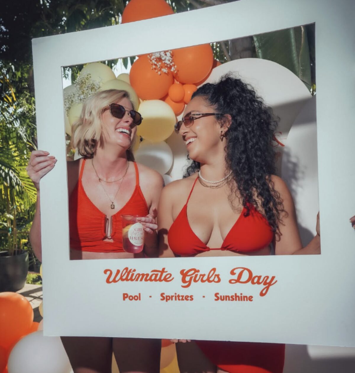 Two friends laughing inside an Ultimate Girls Day photo frame in front of an orange and cream balloon wall