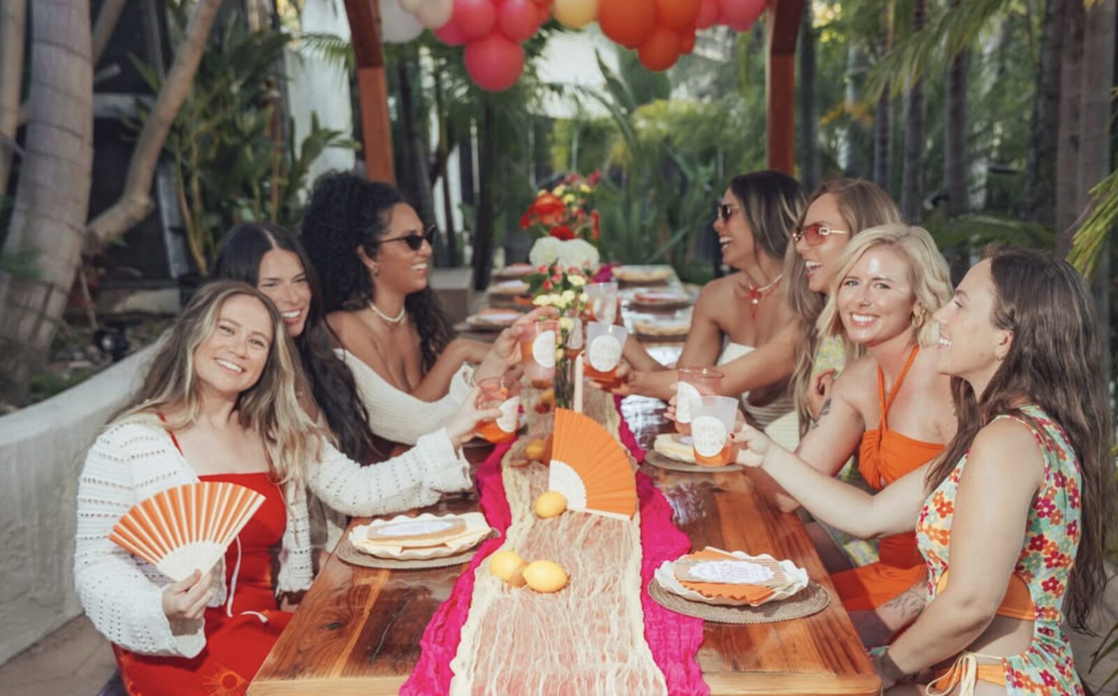 Friends toasting at a styled girls day table with florals, fans and custom cups