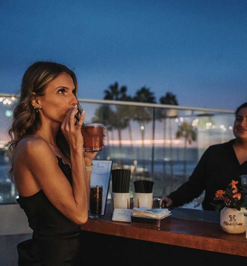 A guest sipping a cocktail at the rooftop bar with palm trees and city lights behind her