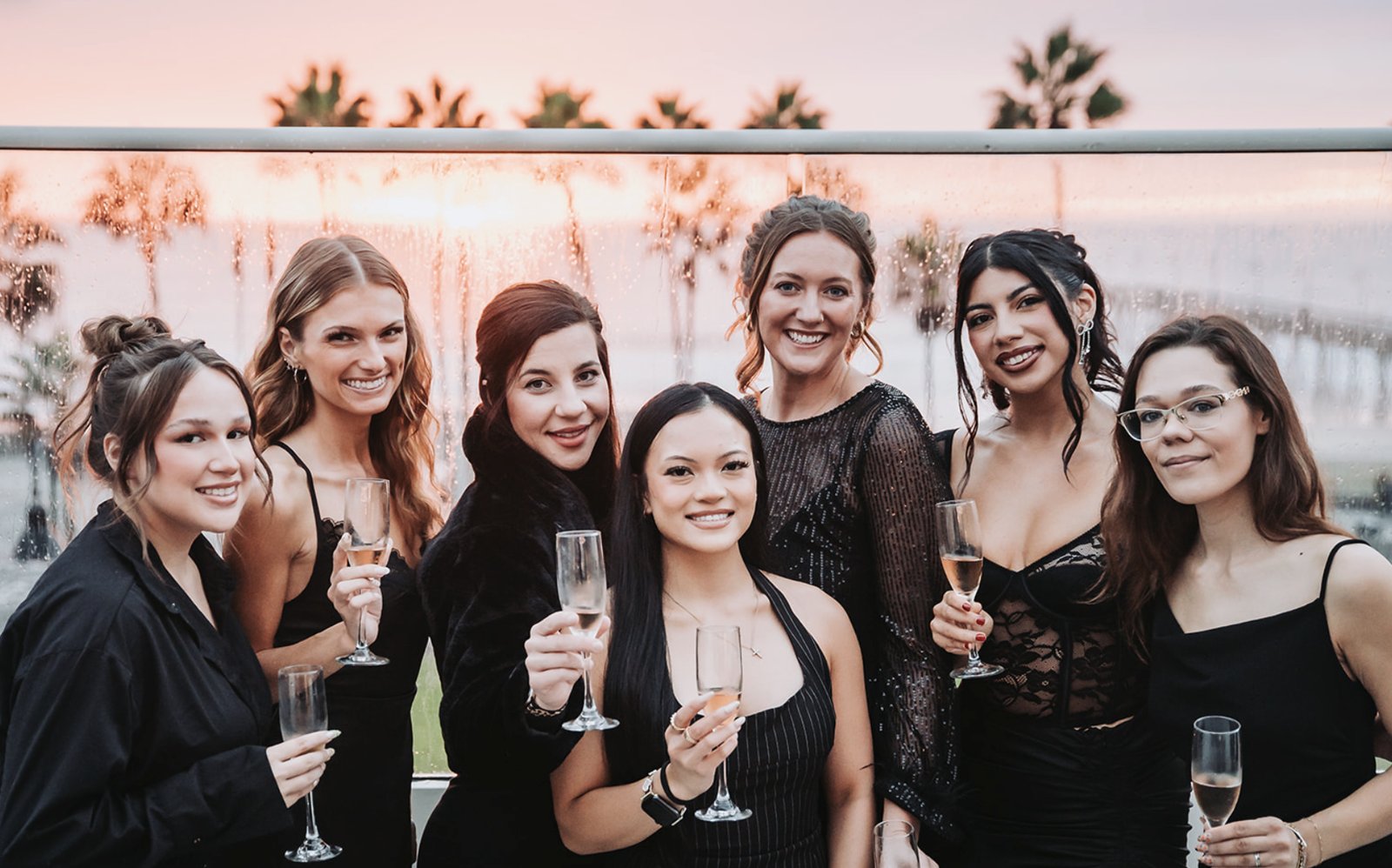 Seven women in black dresses smiling with champagne on the rooftop at sunset