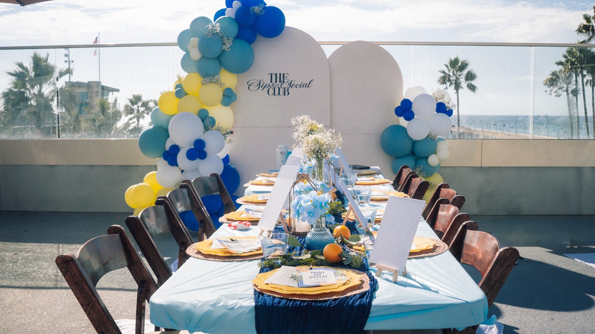 A paint and sip table on a rooftop with a blue and yellow balloon arch and the pier behind