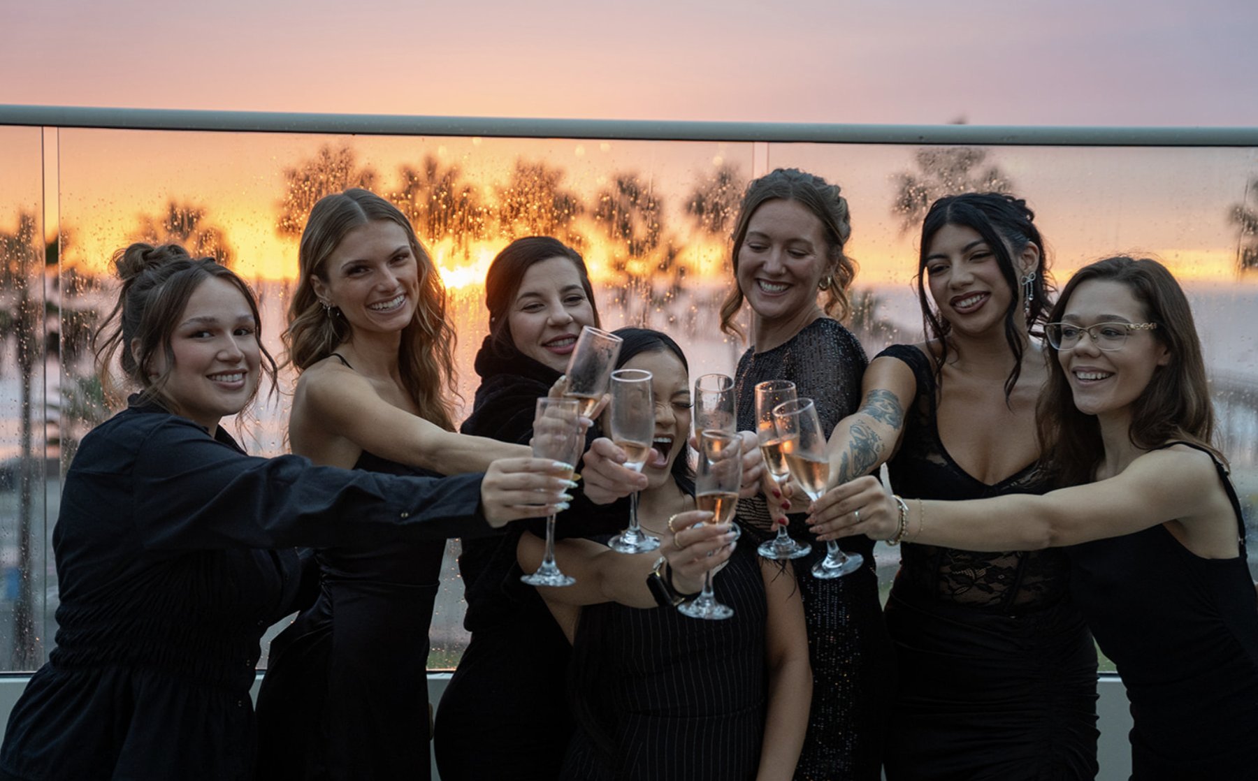 A group of women in black dresses toasting with champagne on the rooftop at sunset, palm trees and the ocean behind them